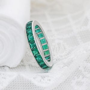 May include: A silver ring with a band of square-cut emerald green gemstones. The ring is set against a white, textured background, showcasing the vibrant green stones and the ring's polished metal.