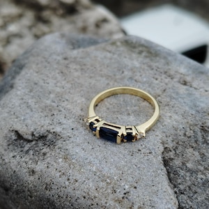 Op de afbeelding: Een gouden ring met een rij blauwe edelstenen en kleine heldere stenen.