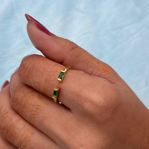 May include: A gold-colored ring with two emerald-green rectangular gemstones. The ring has an open design, with the gemstones set on either side of the opening. The ring is worn on a finger.
