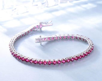 May include: A silver tennis bracelet featuring a continuous line of bright pink gemstones. The bracelet is open, revealing the clasp and the intricate setting of the stones. The background is a pale blue.