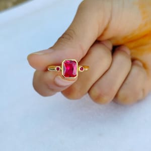 May include: A gold ring featuring a large, rectangular, red gemstone with two smaller red gemstones on either side.