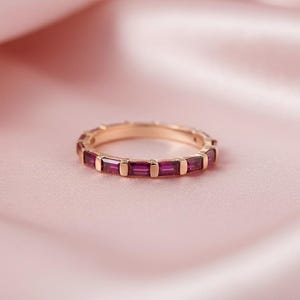 Op de afbeelding: Een roségouden ring met een band van rechthoekige, dieprode edelstenen. De ring is geplaatst tegen een zachte, roze, satijnachtige achtergrond, wat een luxueuze uitstraling creëert. De edelstenen zijn gelijkmatig verdeeld.