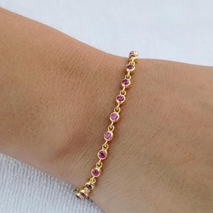 Può includere: Un braccialetto d'oro con una serie di piccole pietre preziose rosa e rotonde. Il braccialetto è delicato e sembra essere fatto d'oro. Le pietre preziose sono uniformemente distanziate lungo il braccialetto.