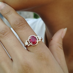 May include: A silver ring featuring a large oval ruby gemstone and two smaller diamond accents.