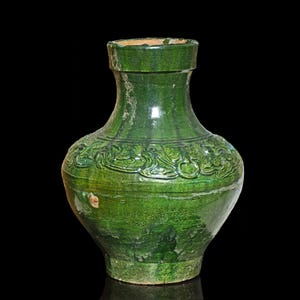 Puede incluir: Un jarrón de cerámica verde con una base ancha y un cuello estrecho. El jarrón tiene un diseño en relieve de flores y hojas estilizadas.