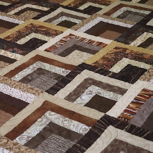 May include: A quilt featuring a repeating log cabin block pattern in various shades of brown, tan, and cream. The fabric squares display diverse textures and prints, including paisley, floral, and geometric motifs, joined by visible stitching.