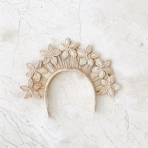 Puede incluir: Una corona floral beige tejida con una base de semicírculo. La corona presenta múltiples flores de cinco pétalos y líneas radiantes, creando un efecto de sol. La diadema está hecha de fibras naturales, ofreciendo una estética bohemia.