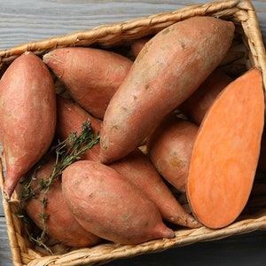 May include: A wicker basket filled with sweet potatoes, some of which are cut in half, with a sprig of thyme on top. A single sweet potato half is on a white and black checked cloth next to a knife.