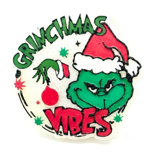 May include: A round, white Christmas ornament featuring the Grinch with a red Santa hat and the words "GRINCHMAS VIBES" in green and red. The Grinch's face is green with a black outline, and the ornament has a festive design.
