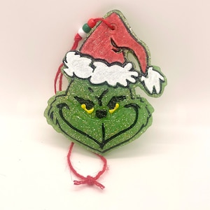 May include: Green Grinch Christmas ornament with a red Santa hat and white trim. The ornament has glitter and a red string for hanging.