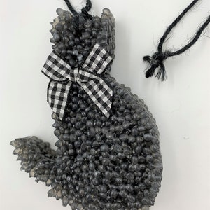 May include: A black cat-shaped air freshener with a gingham bow. The air freshener is made of small, dark gray beads and has a black string for hanging. The bow is black and white.