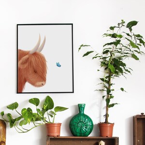 May include: A framed illustration of a brown Highland cow with a blue butterfly flying nearby. The illustration is on a white background and is framed in black. The image is displayed on a white wall with a green plant and a green vase in the foreground.