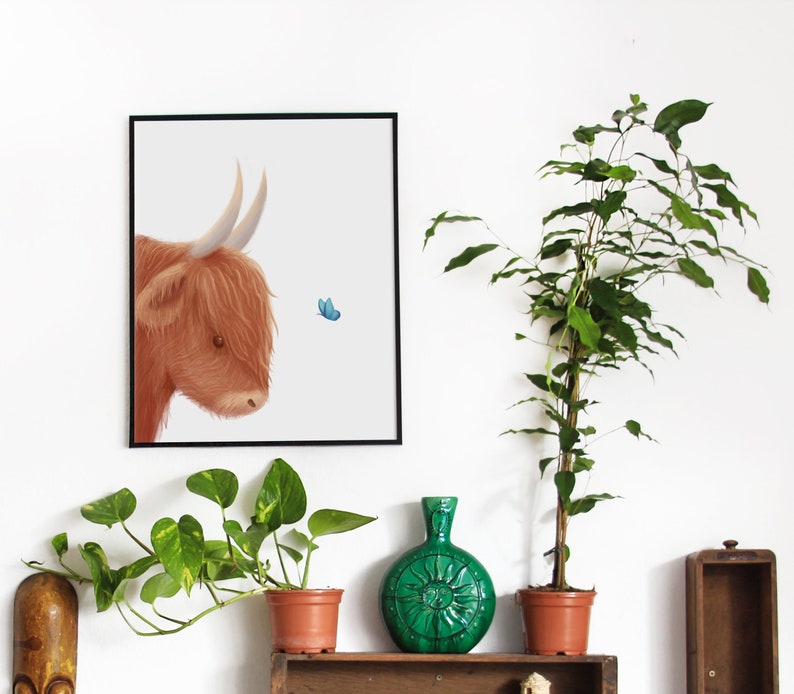 May include: A framed illustration of a brown Highland cow with a blue butterfly flying nearby. The illustration is on a white background and is framed in black. The image is displayed on a white wall with a green plant and a green vase in the foreground.