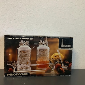 May include: A box for a jam and jelly server set. The set is made of thick acrylic and features a grape design. The box is black with white text that reads "Illusions Thick Acrylic" and "JAM & JELLY SERVER SET". The box also has the company logo "PRODYNE."