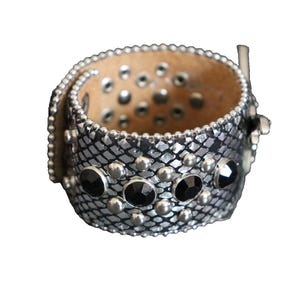 May include: A silver and black cuff bracelet with a snakeskin pattern. The bracelet is adorned with silver studs and black gemstones. The interior of the bracelet is a light brown color.