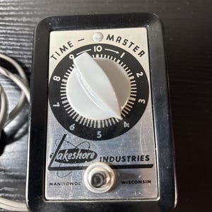 Puede incluir: Un temporizador Time-Master negro y plateado vintage de Lakeshore Industries. El temporizador presenta una esfera blanca con números negros y una perilla gris. El texto "TIME - MASTER" está impreso en la parte superior y "Lakeshore INDUSTRIES" en la inferior.