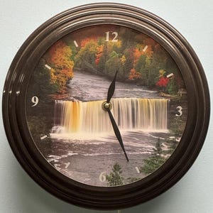 May include: A brown clock with a picture of a waterfall and trees. The clock face has black hands and white numbers. The numbers 12, 9, 3, and 6 are visible.