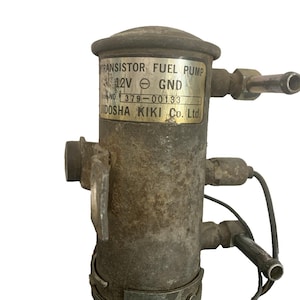 Könnte beinhalten: Eine gebrauchte, verwitterte Kraftstoffpumpe mit dem Text "TRANSISTOR FUEL PUMP 12V GND" und "UIDOSHA KIKI Co. Ltd." auf einer Metallplatte. Die Pumpe hat mehrere Metallrohre und ein schwarzes Kabel.