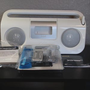 May include: A white portable radio with a built-in handle, two circular speakers, and a central silver control panel. Included are a small remote control in plastic packaging, a product registration card, and an XpressEZ user manual.
