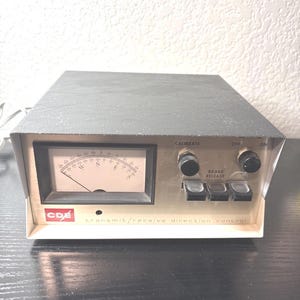 Op de afbeelding: Een vintage CDE zend/ontvangst richting controle-eenheid. Het apparaat heeft een grijze bovenkant en een beige voorpaneel met een meter, knoppen en knoppen. De tekst "CALIBRATE", "OFF", "ON" en "BRAKE RELEASE" is zichtbaar.