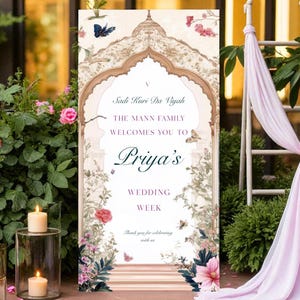 May include: A decorative wedding sign with floral and butterfly accents. The sign features text that reads "Priya's Wedding Week". The sign is framed by an archway design and surrounded by greenery and candles.