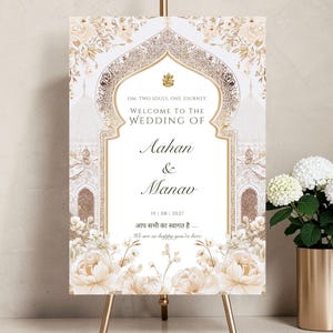 May include: A wedding welcome sign on a gold easel. The sign features floral and architectural designs in cream and gold. Text on the sign reads "Welcome to the Wedding of Aahan & Manav" and includes a date. A gold vase with white flowers is to the right.