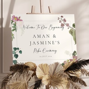 May include: A wedding welcome sign on an easel, featuring floral accents and the text "Welcome To Our Beginning" with names and date. The sign is decorated with dried flowers and greenery.
