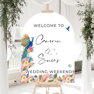 May include: Welcome to Cameron & Sonia's Wedding Weekend sign with a peacock and floral border. The sign is white with black text and is displayed on an easel.