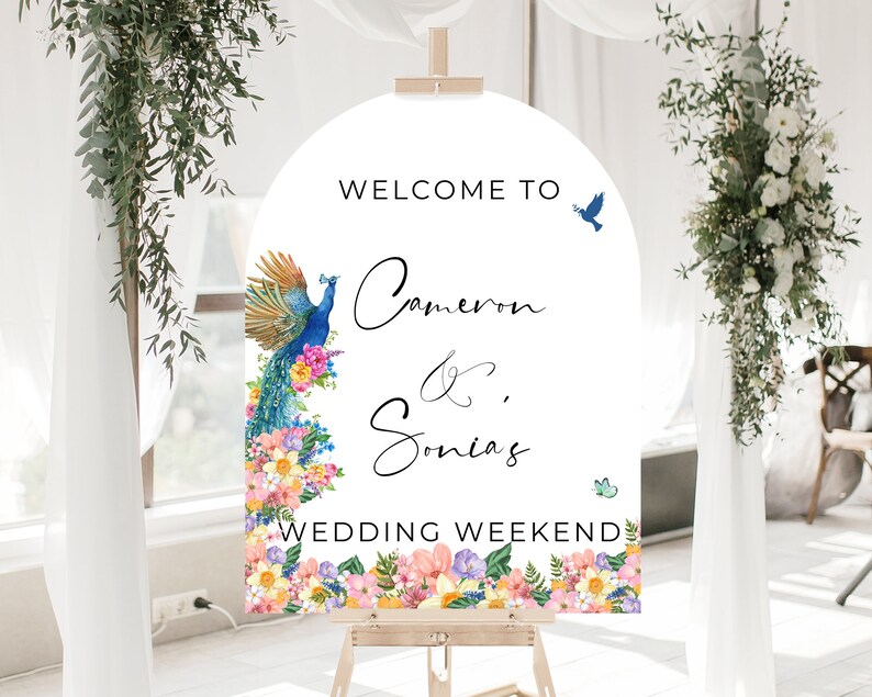 May include: Welcome to Cameron & Sonia's Wedding Weekend sign with a peacock and floral border. The sign is white with black text and is displayed on an easel.