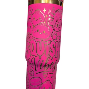 Puede incluir: Un vaso de color rosa brillante con tapa transparente y borde dorado, con dibujos de líneas doradas de dumplings, una vaca de las Tierras Altas y flores. El texto "SQUISH VIBES" aparece en una fuente llamativa y estilizada.
