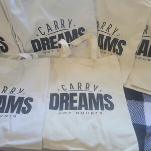 Op de afbeelding: Vier witte draagtassen met zwarte tekst. De tekst op de tassen luidt "Carry Dreams Not Doubts".