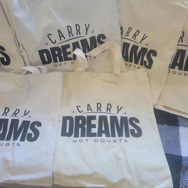 Op de afbeelding: Vier witte draagtassen met zwarte tekst. De tekst op de tassen luidt "Carry Dreams Not Doubts".