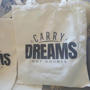 Op de afbeelding: Twee witte canvas tassen met zwarte handvatten. De tas aan de rechterkant heeft de tekst "Carry Dreams Not Doubts" erop gedrukt.