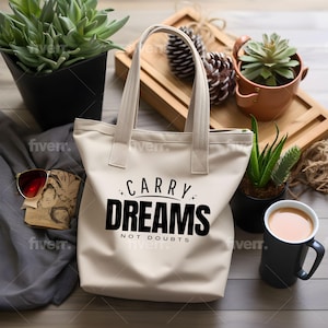 Op de afbeelding: Een witte canvas tote bag met zwarte tekst die "Carry Dreams Not Doubts" zegt. De tas ligt op een houten oppervlak met een kop koffie en planten op de achtergrond.