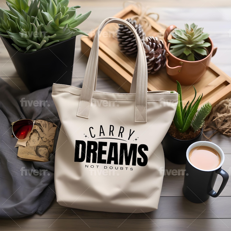 Op de afbeelding: Een witte canvas tote bag met zwarte tekst die "Carry Dreams Not Doubts" zegt. De tas ligt op een houten oppervlak met een kop koffie en planten op de achtergrond.