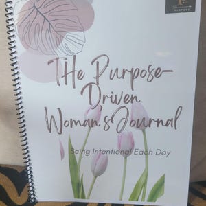 K&ouml;nnte beinhalten: Ein wei&szlig;es Spiral-Notizbuch mit einem rosa-braunen Blumendesign. Der Umschlag tr&auml;gt die Aufschrift "The Purpose-Driven Woman's Journal" und "Being Intentional Each Day".