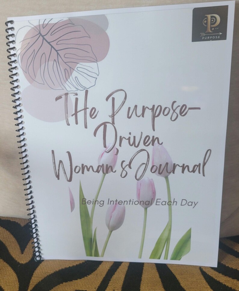 K&ouml;nnte beinhalten: Ein wei&szlig;es Spiral-Notizbuch mit einem rosa-braunen Blumendesign. Der Umschlag tr&auml;gt die Aufschrift "The Purpose-Driven Woman's Journal" und "Being Intentional Each Day".