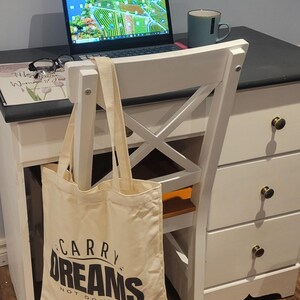 Op de afbeelding: Een witte canvas draagtas met zwarte tekst die "Carry Dreams Not Doubts" luidt. De tas hangt aan de rugleuning van een witte stoel met een houten zitting. Een laptop en een mok staan op een wit bureau met zwarte lades.