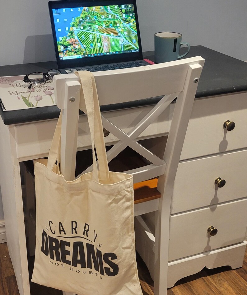 Op de afbeelding: Een witte canvas draagtas met zwarte tekst die "Carry Dreams Not Doubts" luidt. De tas hangt aan de rugleuning van een witte stoel met een houten zitting. Een laptop en een mok staan op een wit bureau met zwarte lades.
