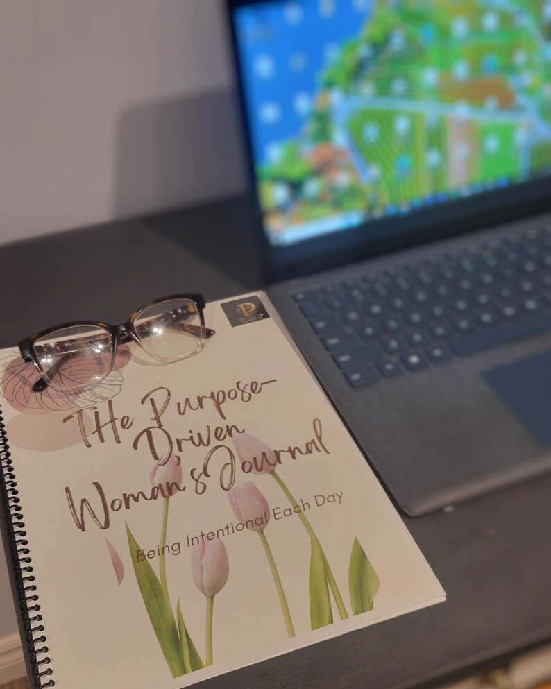 K&ouml;nnte beinhalten: Ein geschlossenes Spiralbuch mit einem hellbraunen Einband und dem Text "The Purpose-Driven Woman's Journal" und "Being Intentional Each Day". Der Einband zeigt eine Aquarell-Illustration von rosa Tulpen mit gr&uuml;nen Bl&auml;ttern.
