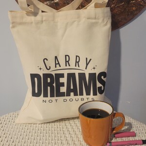 Op de afbeelding: Een witte canvas draagtas met zwarte tekst die "Carry Dreams Not Doubts" zegt. De tas ligt op een wit rieten oppervlak met een bruine koffiemok en roze pennen.