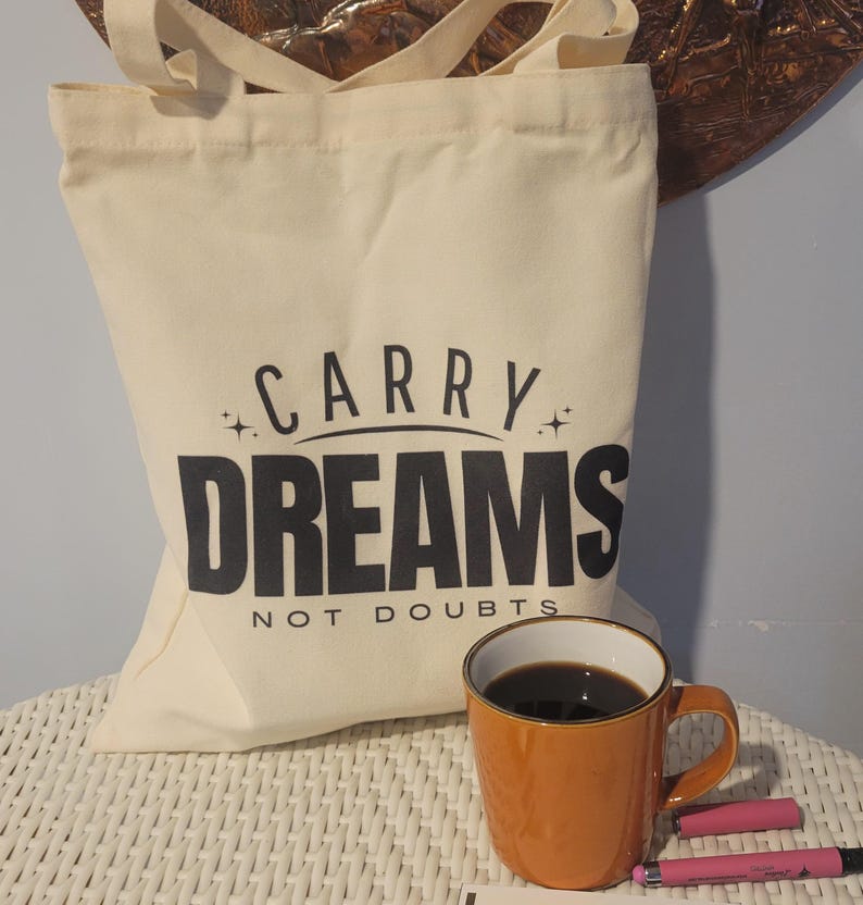 Op de afbeelding: Een witte canvas draagtas met zwarte tekst die "Carry Dreams Not Doubts" zegt. De tas ligt op een wit rieten oppervlak met een bruine koffiemok en roze pennen.