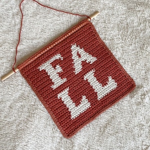 May include: A crocheted wall hanging with the word "FALL" in white yarn on a rust-colored background. The hanging is attached to a wooden dowel with a string loop.