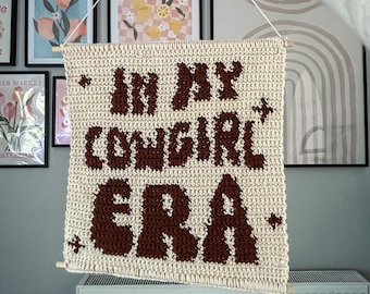 May include: Cream-colored, crocheted wall hanging with the text "IN MY COWGIRL ERA" in brown. The banner is suspended by a wooden dowel and string. The background includes framed art and a radiator.