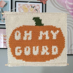 May include: A cream-coloured, crocheted wall hanging with an orange pumpkin design and the words "OH MY GOURD" in white. The pumpkin has a green stem. Wooden dowels are at the top and bottom. A seasonal, decorative item.