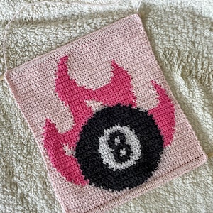 Puede incluir: Un tapiz de pared hecho a mano, tejido a ganchillo, con un fondo rosa y un diseño de bola ocho en blanco y negro, rodeado de llamas rosas. La pieza es cuadrada y tiene un lazo para colgar.