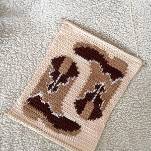 Puede incluir: Un tapiz de pared de ganchillo beige, marr&oacute;n y blanco. La pieza rectangular presenta un patr&oacute;n repetido de formas abstractas en diferentes tonos de marr&oacute;n y blanco sobre un fondo beige. La parte superior tiene clavijas de madera y una cuerda para colgar.