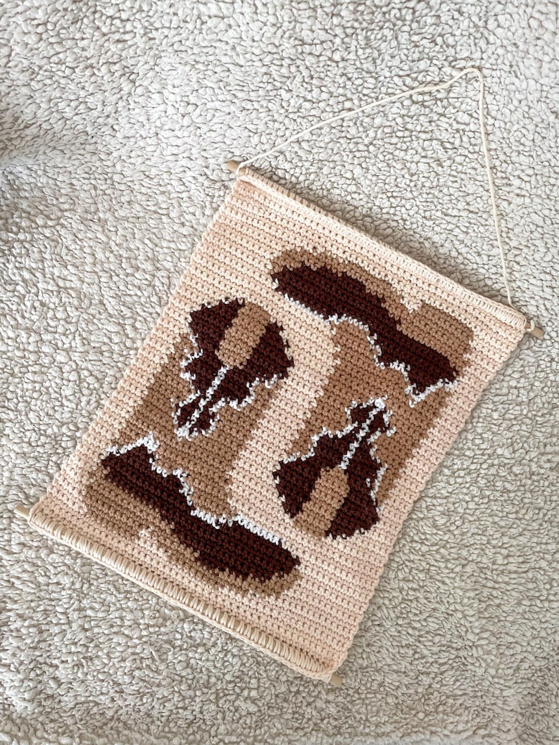 Puede incluir: Un tapiz de pared de ganchillo beige, marr&oacute;n y blanco. La pieza rectangular presenta un patr&oacute;n repetido de formas abstractas en diferentes tonos de marr&oacute;n y blanco sobre un fondo beige. La parte superior tiene clavijas de madera y una cuerda para colgar.