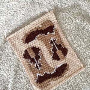 Puede incluir: Un tapiz de pared tejido a ganchillo en beige y marr&oacute;n con un dise&ntilde;o geom&eacute;trico. La pieza rectangular presenta un fondo beige claro con detalles en marr&oacute;n oscuro y blanco. Una cuerda color crema est&aacute; unida para colgar.