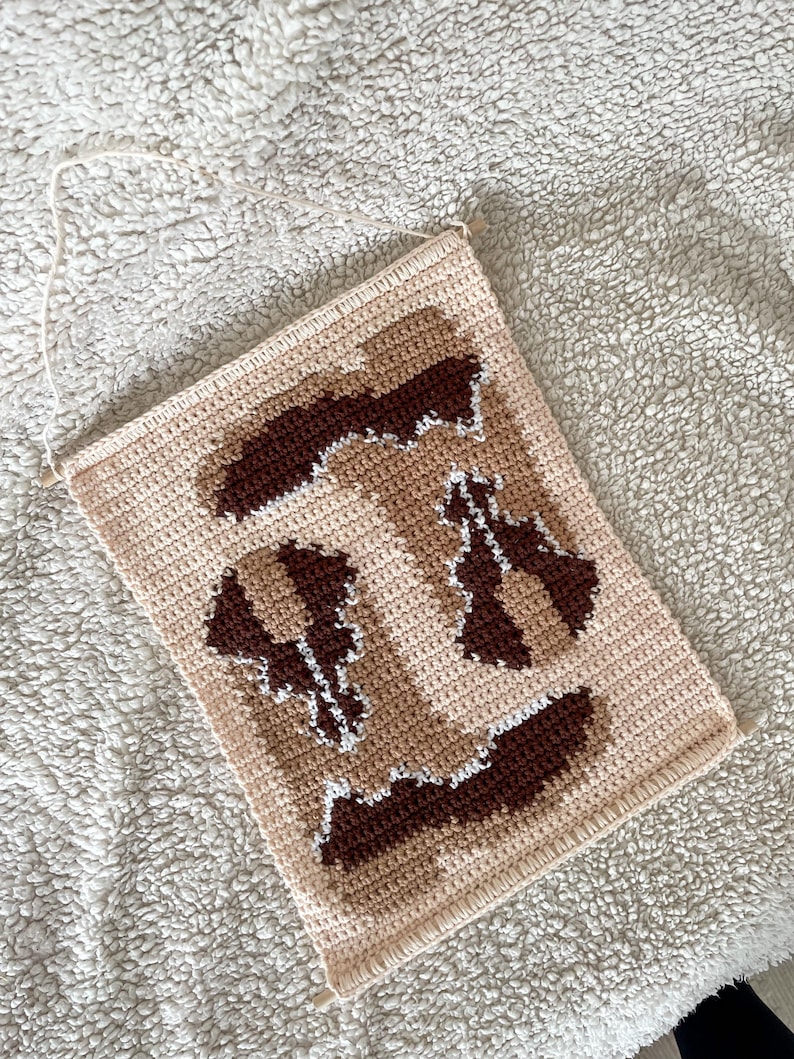 Puede incluir: Un tapiz de pared tejido a ganchillo en beige y marr&oacute;n con un dise&ntilde;o geom&eacute;trico. La pieza rectangular presenta un fondo beige claro con detalles en marr&oacute;n oscuro y blanco. Una cuerda color crema est&aacute; unida para colgar.